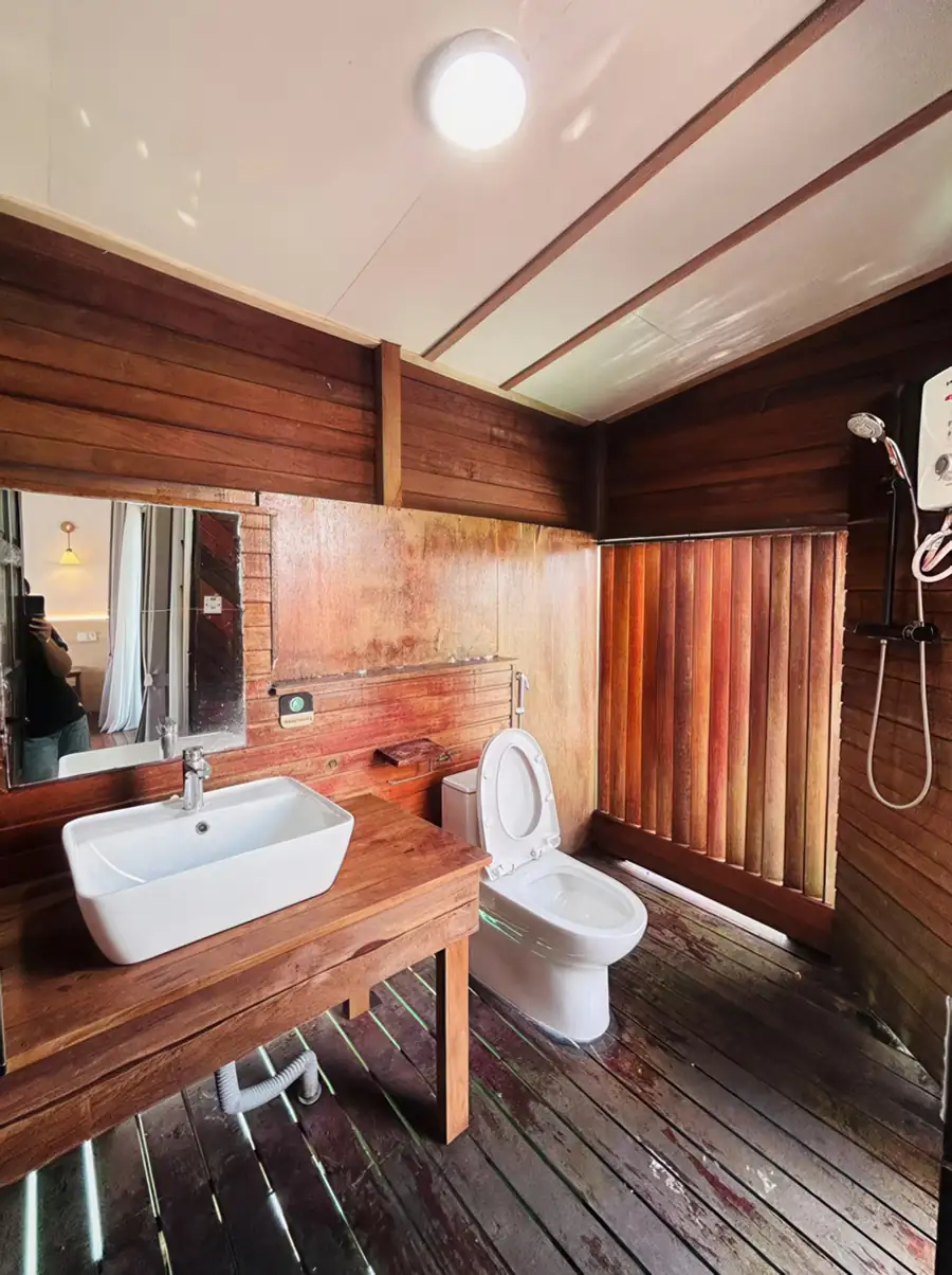 A practical and clean bathroom showing a classic white washbasin, a wall mirror, a ceramic toilet, light-colored tiled walls, and a corner electric shower space at Sea Coral Resort.