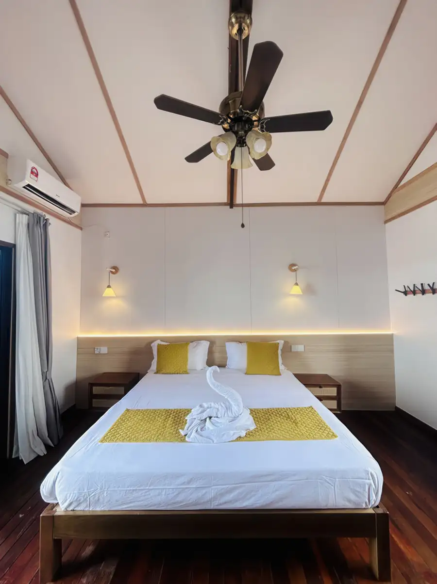 An inviting guest room at Sea Coral Resort near Semporna showcasing a spacious bed, minimalist decor, warm wooden plank floors with varnished timber, ceiling fan, and air conditioning.