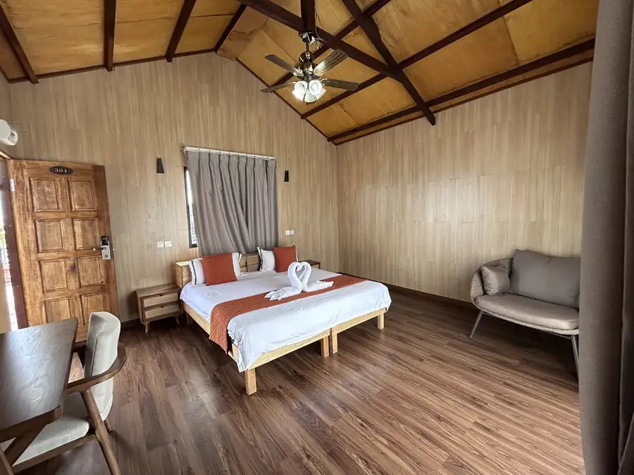 A bright bedroom at Sea Coral Resort near Pulau Bum Bum featuring a large bed, a comfortable seating area, authentic wooden walls with glossy hardwood floors, and AC.
