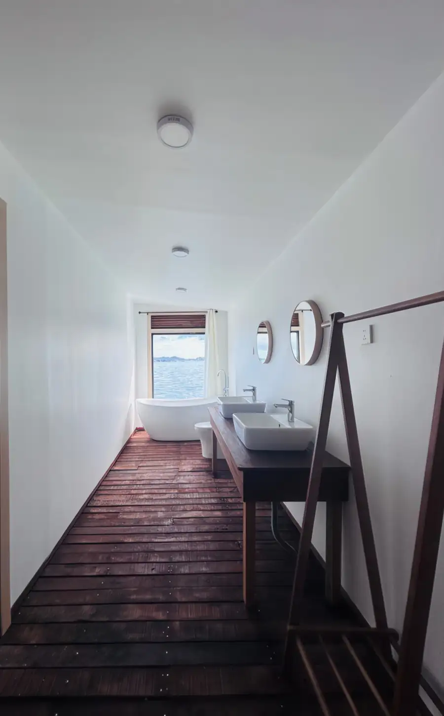 A long, airy bathroom at Sea Coral Resort featuring white walls, wood plank floors, a freestanding tub, and a wooden vanity with sinks leading to an ocean view window.