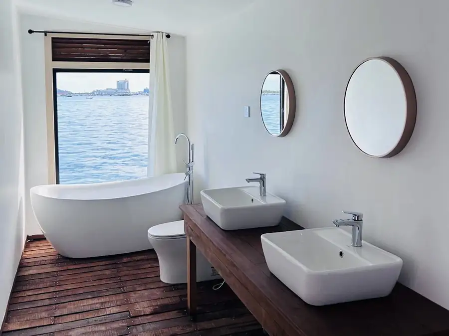 A clean and practical bathroom at Sea Coral Resort showcasing a classic pedestal washbasin, a wall mirror, a ceramic toilet, patterned floor tiles, and a dedicated corner space fitted with an electric shower.