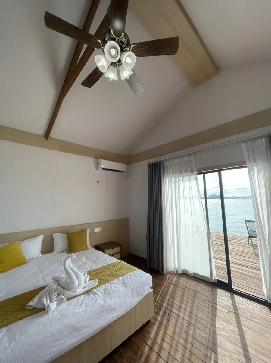 A relaxing bedroom sanctuary at Sea Coral Resort near Semporna offering a large bed, panoramic glass doors opening onto the balcony, ceiling fan, smooth varnished timber flooring, and A/C.
